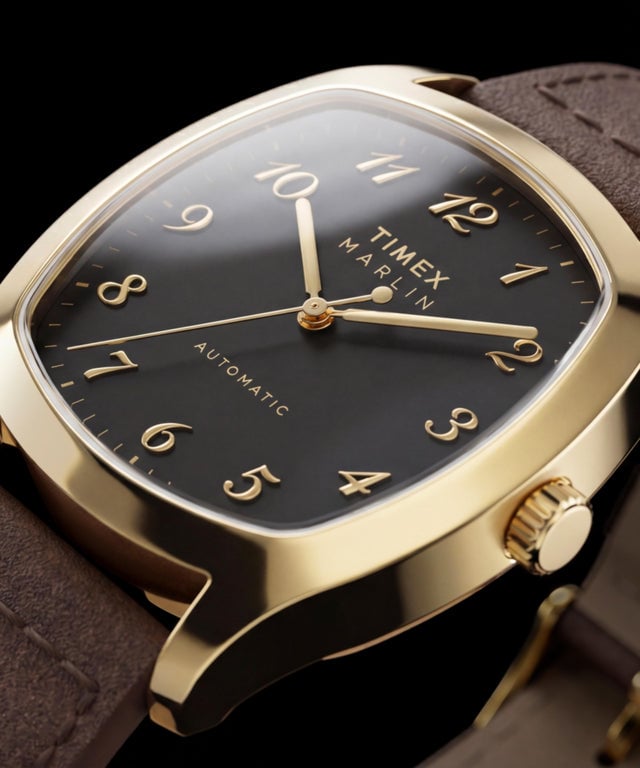 Close-up do Timex Marlin Chatham Automatic em tom dourado, mostrando o mostrador preto, os algarismos arábicos aplicados em tom dourado, os ponteiros em tom dourado e a pulseira de couro castanho