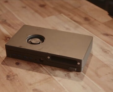 Um console de videogame preto e compacto, com um leitor de discos integrado, portas USB e um ventilador de resfriamento na parte superior, apoiado sobre uma mesa de madeira.