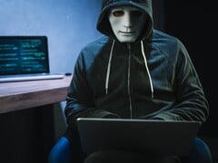 Uma figura mascarada e encapuzada trabalha em um laptop em uma sala escura.