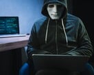 Uma figura mascarada e encapuzada trabalha em um laptop em uma sala escura.