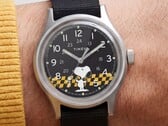 Close-up do mostrador preto do Timex MK1, exibindo o Snoopy contra um padrão xadrez amarelo e preto, com marcadores de horas em algarismos arábicos e ponteiros luminosos.
