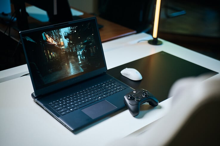 Um notebook para jogos Alienware aberto, instalado sobre uma mesa branca de estúdio, exibindo uma cena de rua escura e reflexiva com detalhes nítidos obtidos por rastreamento de trajetória, acompanhado por um controle preto e um mouse sem fio branco.