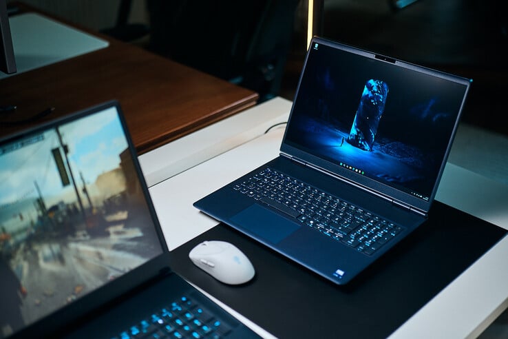 Um laptop para jogos de gama média, na cor azul escuro (Alienware Aurora 16X), inclinado sobre um tapete de mesa com um papel de parede de ficção científica azul brilhante, posicionado ao lado de um mouse sem fio branco, com outro laptop desfocado em primeiro plano.