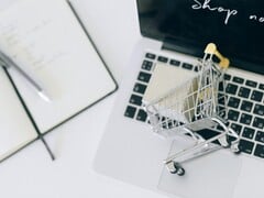 Carrinho de compras em miniatura no teclado de um laptop, com a mensagem “Compre agora” na tela