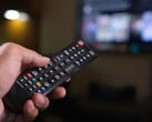 Mão segurando um controle remoto com uma TV desfocada ao fundo