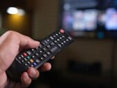 Mão segurando um controle remoto com uma TV desfocada ao fundo