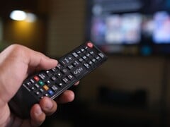 Mão segurando um controle remoto com uma TV desfocada ao fundo