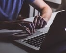 Mãos digitando no teclado de um MacBook sob luz fraca, com o logotipo da Apple brilhando
