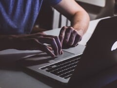 Mãos digitando no teclado de um MacBook sob luz fraca, com o logotipo da Apple brilhando
