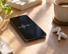 Smartphone exibindo uma forma de onda de áudio ao lado de fones de ouvido sem fio e uma xícara de café sobre uma mesa de madeira