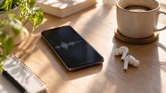 Smartphone exibindo uma forma de onda de áudio ao lado de fones de ouvido sem fio e uma xícara de café sobre uma mesa de madeira