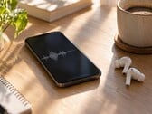 Smartphone exibindo uma forma de onda de áudio ao lado de fones de ouvido sem fio e uma xícara de café sobre uma mesa de madeira