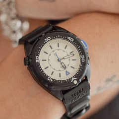 O Timex Intrepid x Dimepiece Blackout Edition apresenta uma caixa em aço inoxidável preto, mostrador creme e detalhes roxos