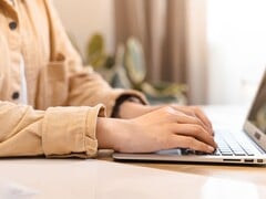 Mãos digitando no teclado de um laptop aberto sobre uma mesa bem iluminada