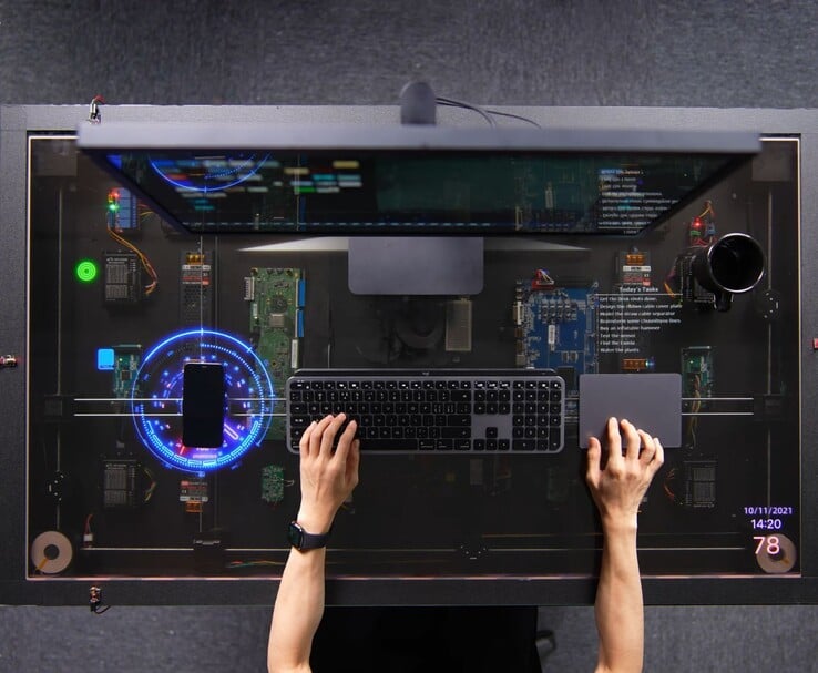 A mesa inspirada no AirPower, criada pelo HTX Studio, em uso, com um celular carregando no visor transparente ao lado de um teclado, trackpad e monitor.