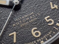 Close-up do mostrador preto texturizado do Timex Expedition Field Post Titanium, com algarismos arábicos, ponteiros e marcadores luminosos e janela de data.