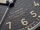 Close-up do mostrador preto texturizado do Timex Expedition Field Post Titanium, com algarismos arábicos, ponteiros e marcadores luminosos e janela de data.