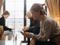 Uma adolescente olha para o smartphone, com dois adultos sentados desfocados ao fundo.