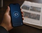 Uma mão segura um smartphone que exibe o ícone de um chatbot; ao lado dele, encontra-se um jornal impresso.
