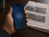 Uma mão segura um smartphone que exibe o ícone de um chatbot; ao lado dele, encontra-se um jornal impresso.