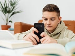 Adolescente vestindo um moletom com capuz digitando em um smartphone, com livros escolares abertos à sua frente