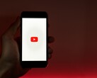 Uma mão segurando um smartphone com o logotipo do YouTube iluminado na tela, tendo como pano de fundo uma cor vermelha