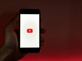 Uma mão segurando um smartphone com o logotipo do YouTube iluminado na tela, tendo como pano de fundo uma cor vermelha