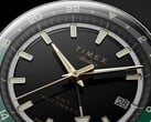 Grande plano do mostrador preto do Timex Atelier GMT Première M1a, com ponteiros em tom dourado, marcadores de hora luminescentes, janela de data e anel GMT de 24 horas preto e verde.