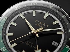 Grande plano do mostrador preto do Timex Atelier GMT Première M1a, com ponteiros em tom dourado, marcadores de hora luminescentes, janela de data e anel GMT de 24 horas preto e verde.