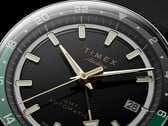Grande plano do mostrador preto do Timex Atelier GMT Première M1a, com ponteiros em tom dourado, marcadores de hora luminescentes, janela de data e anel GMT de 24 horas preto e verde.