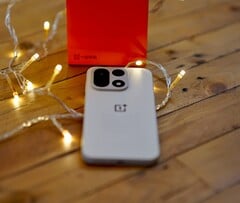 Um OnePlus 15 está sobre uma mesa de madeira, rodeado por luzes de corda que criam um ambiente aconchegante, com uma caixa de varejo da OnePlus em vermelho vivo posicionada ao fundo.