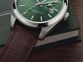 Close-up do relógio Timex Legacy Day and Date, mostrando seu mostrador verde com acabamento raios de sol, aro em tom prateado e asas escovadas, coroa estriada e pulseira de couro marrom texturizado, diante de uma caixa verde-escura e sobre uma superfície de madeira.
