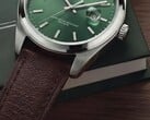 Close-up do relógio Timex Legacy Day and Date, mostrando seu mostrador verde com acabamento raios de sol, aro em tom prateado e asas escovadas, coroa estriada e pulseira de couro marrom texturizado, diante de uma caixa verde-escura e sobre uma superfície de madeira.