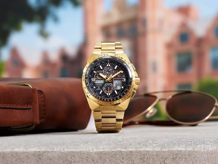 Relógio de piloto Citizen Promaster Skyhawk A-T JY8144-50E em tom dourado, exposto na vertical sobre uma prateleira ao lado de uma bolsa de couro marrom e óculos de sol, com uma paisagem urbana desfocada ao fundo.
