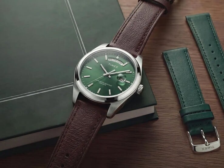 O Timex Legacy Day and Date com seu mostrador verde e pulseira de couro marrom, exibido sobre um livro verde-escuro ao lado da pulseira intercambiável de couro verde.