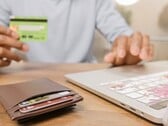Pessoa segurando um cartão bancário em frente a um laptop, com uma carteira sobre a mesa