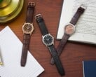 Três relógios Citizen Rolan dispostos sobre uma mesa de madeira ao lado de um caderno, uma caneta, um abajur e um livro aberto, incluindo o modelo AW2110-14Z com mostrador cor de salmão.