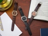 Três relógios Citizen Rolan dispostos sobre uma mesa de madeira ao lado de um caderno, uma caneta, um abajur e um livro aberto, incluindo o modelo AW2110-14Z com mostrador cor de salmão.