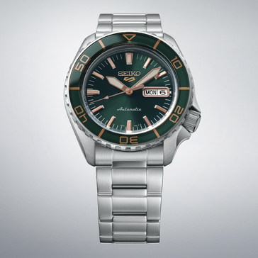 Seiko 5 Sports SKX HDB005 com mostrador verde-escuro, luneta verde a condizer, ponteiros e índices em tom de ouro rosa e bracelete de aço inoxidável, apresentado contra um fundo cinzento-claro.