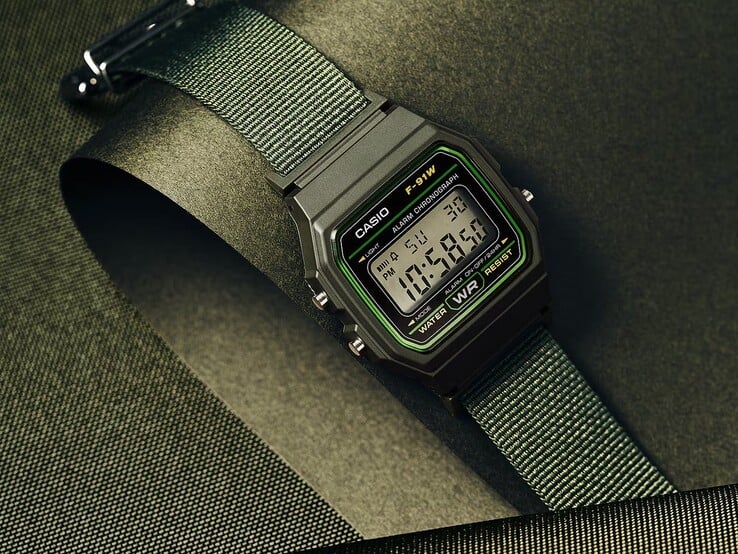 Relógio digital Casio F-91WF-3A com caixa preta com detalhes em verde e pulseira de tecido verde trançado, fotografado contra um fundo texturizado verde-escuro.