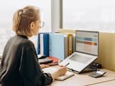Mulher de óculos trabalha em uma planilha no laptop, com pastas e uma calculadora sobre a mesa