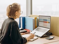 Mulher de óculos trabalha em uma planilha no laptop, com pastas e uma calculadora sobre a mesa