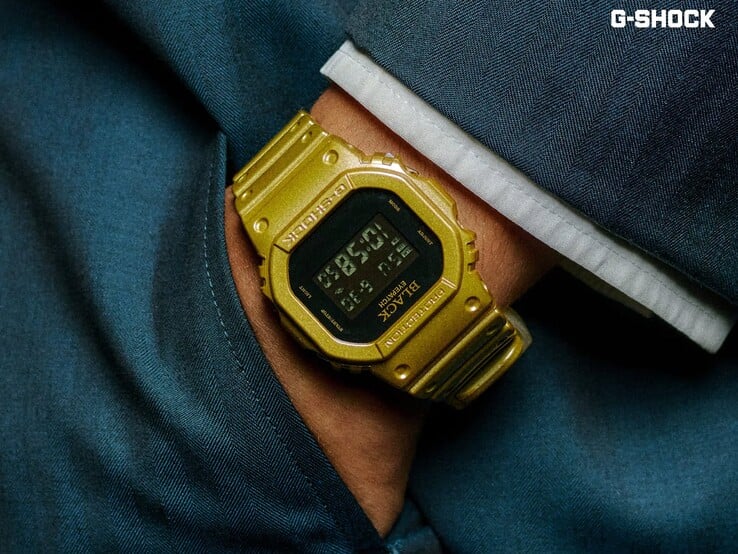 Close-up de um relógio Casio G-Shock DW-5600BEP-9 dourado usado no pulso contra um tecido azul escuro. O relógio possui um visor digital preto, caixa e pulseira douradas, além da marca BlackEyePatch no bisel e no mostrador.