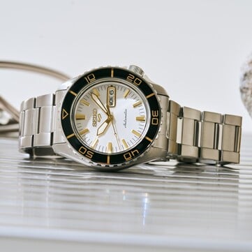 Relógio Seiko 5 Sports SKX HDB004 com mostrador branco-prateado, luneta giratória preta com marcações douradas, ponteiros e índices em tom dourado e bracelete de aço inoxidável, fotografado em ângulo.