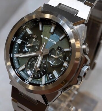 Close-up do Casio G-Shock GST-W1000D-3A com mostrador verde, mostrando seu mostrador com textura geométrica, três submostradores, marcadores luminosos e a luneta em aço inoxidável polido.