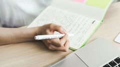 Uma pessoa está escrevendo anotações em um caderno ao lado de um laptop aberto