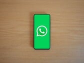 Smartphone sobre uma mesa de madeira, exibindo a tela inicial verde do WhatsApp com seu logotipo.