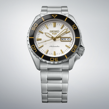 Seiko 5 Sports SKX HDB004 com mostrador branco-prateado, luneta preta com marcações douradas, ponteiros e índices em tom dourado e bracelete de aço inoxidável, apresentado sobre um fundo cinza-claro.